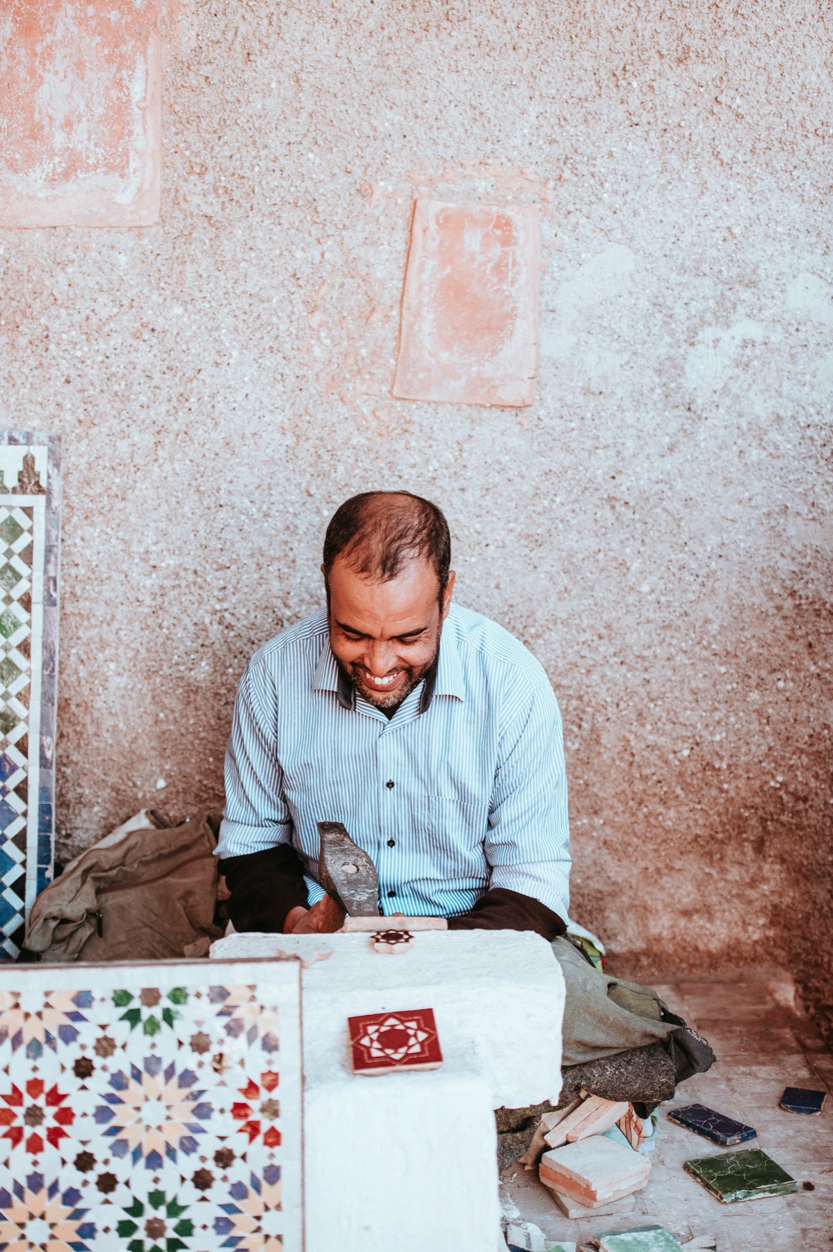 A smiling zellige maâlem hand-cutting a glazed tile with a chisel hammer