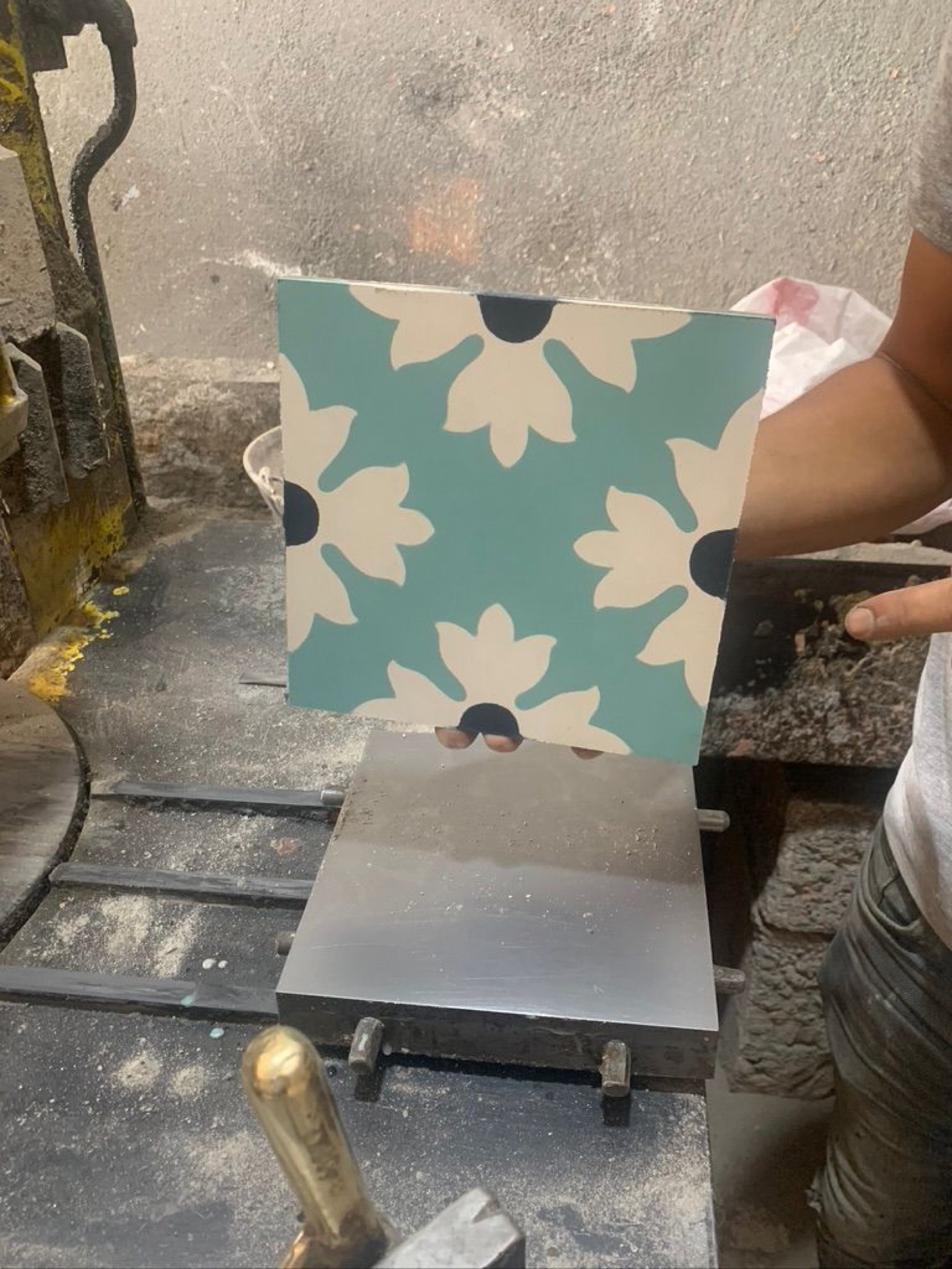 A floral-pattern cement tile lifted from the press, held against the workshop floor